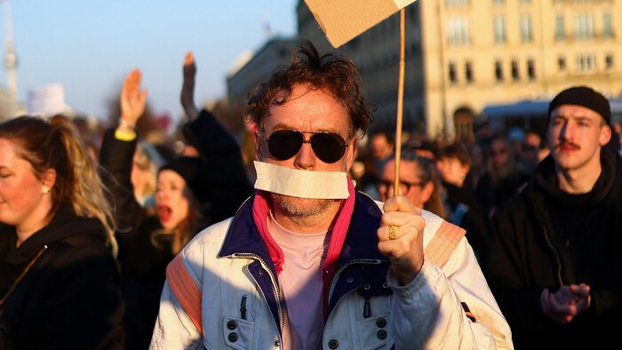 Un homme avec un ruban sur la bouche tient une pancarte pendant une manifestation.