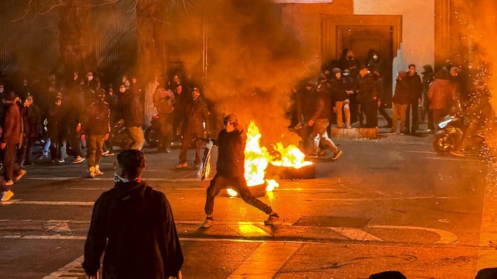 Des Iraniens se rassemblent tout en bloquant une rue lors d'une manifestation. Un objet est incendié au milieu de la rue tandis qu'un manifestant lance un objet.