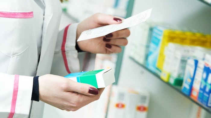 Une pharmacienne en blouse blanche tenant une boîte de médicament dans une main et une ordonnance dans l’autre.