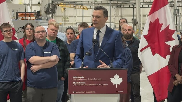 Evan Solomon, debout derrière un lutrin, est entouré de travailleurs d'une usine.