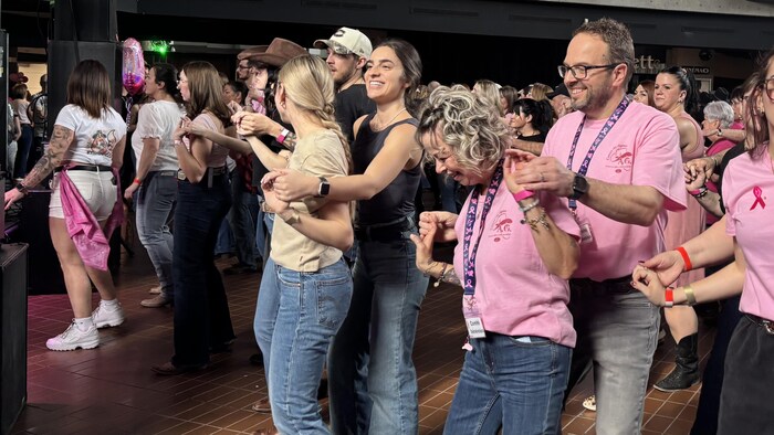 Des participants de tous âges dansent ensemble en se tenant par les mains ou par les bras. La majorité porte des t-shirts roses, certains avec des rubans de sensibilisation. L'action se déroule dans une salle spacieuse avec une mezzanine.