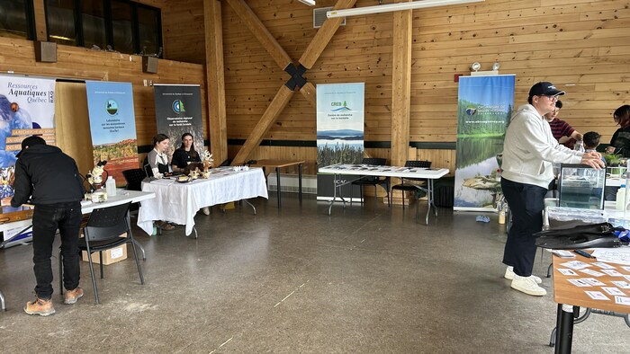 Cette photographie montre une salle en bois abritant plusieurs stands d'information sur l'environnement et l'eau au Québec. Des intervenants et des visiteurs s'activent autour de tables couvertes de nappes blanches, de dépliants, de microscopes et d'affiches promotionnelles.