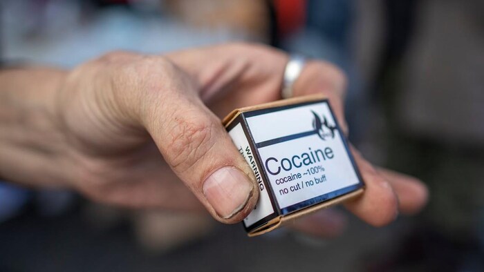 A hand holds a small box with an inscription that reads "Cocaine - 100% - no cut/no buff",