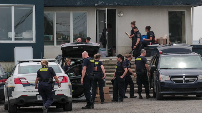 Devant un immeuble, plusieurs personnes vêtues de gilets pare-balles et d'armes embarquent des boîtes dans plusieurs voitures.