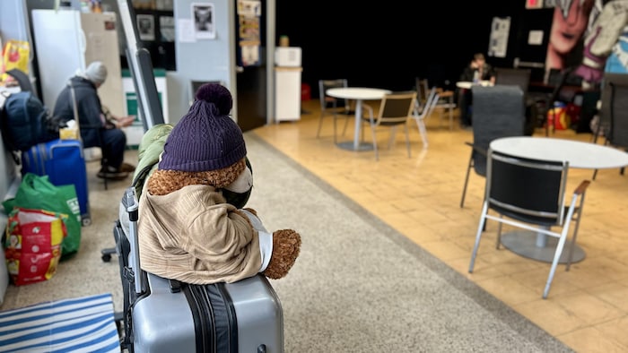 Un homme assis au loin sur une chaise. Devant lui, une valise avec un nounours.