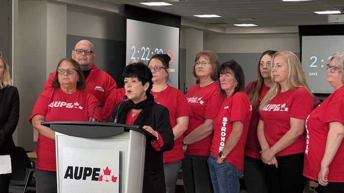 Des personnes habillées en rouge se tiennent derrière une femme parlant devant un lutrin.