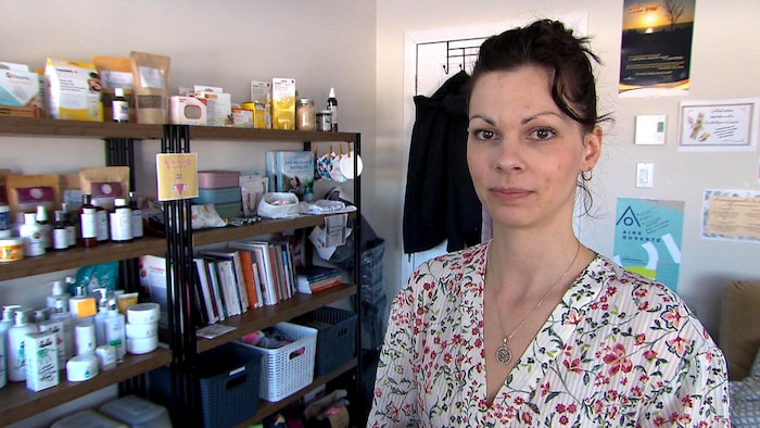 Une femme regarde la caméra. Une étagère garnie de livres et de produits est visible à sa droite.