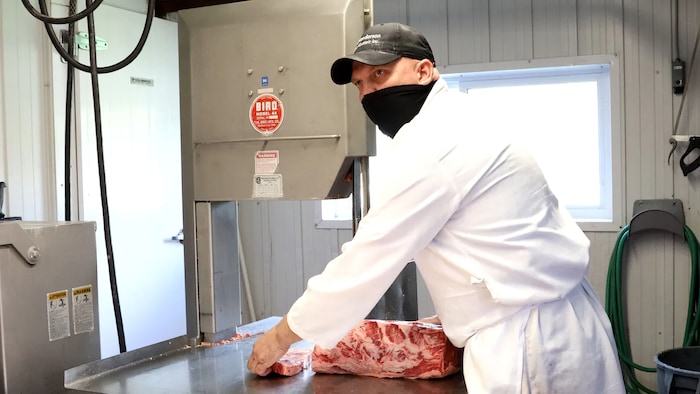 Un boucher vêtu de blanc, passe un bloc viande dans une scie pour en faire des tranches épaisses.