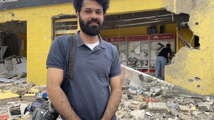 Un homme portant une caméra pose devant un bâtiment détruit.