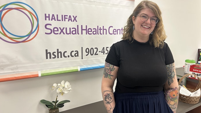 Abbey Ferguson pose pour une photo dans une clinique médicale, devant un bureau et une bannière qui identifie le centre de santé sexuelle.
