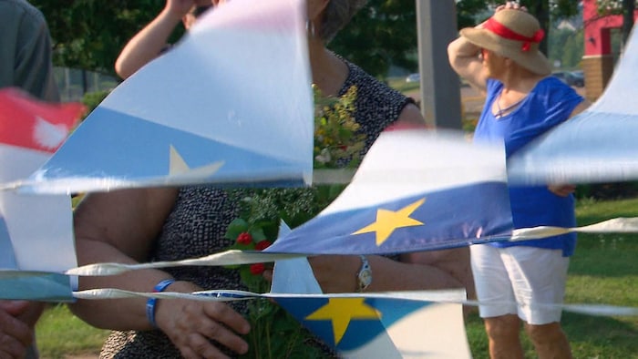 De petits drapeaux de l'Acadie sur une corde voltigent dans le vent. Des gens sont autour, dans un parc.
