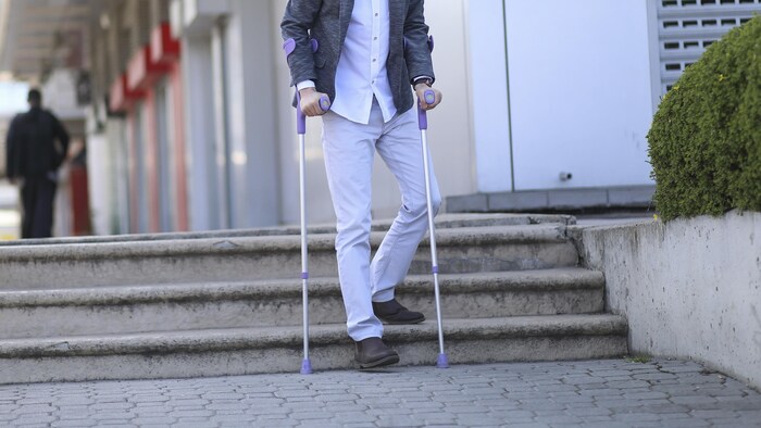 Un homme dans la vingtaine qui marche avec deux cannes descend les trois marches d'un escalier, devant un édifice.