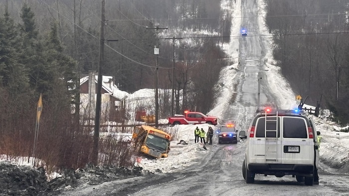 Des véhicules d'urgence et des membres d'équipes d'urgence se trouvent près d'un autobus scolaire accidenté qui gît au bas d'une grande côte. La chaussée est glacée. De la neige et de la gadoue recouvrent les fossés, dont celui où se trouve le véhicule accidenté.