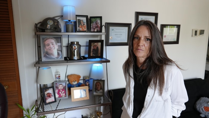 Une photo de Christine D'Astous dans son salon dans son appartement. L'urne et des photos de son fils sont installées en arrière.