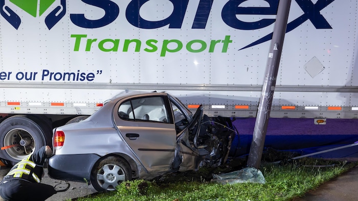Un travailleur est accroupi derrière une petite voiture écrasée sous la remorque d’un camion de l’entreprise Safex Transport. Il s'affaire à mettre un crochet pour pouvoir la remorquer. 