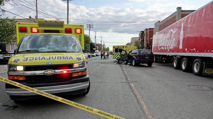 Derrière un ruban de balisage, deux ambulances sont à proximité d'un véhicule accidenté, arrêté au milieu d'une rue, à côté d'un camion-remorque de Coca Cola.