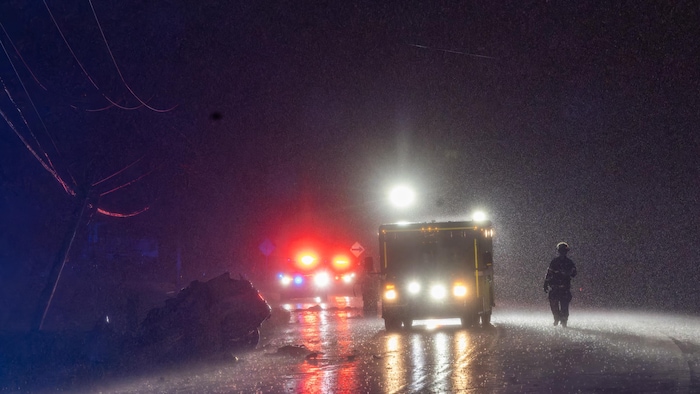 De nuit, une voiture engouffrée dans un poteau. Les gyrophares des véhicules d'urgence éblouissent en reflétant dans la pluie. Un pompier marche vers la scène.