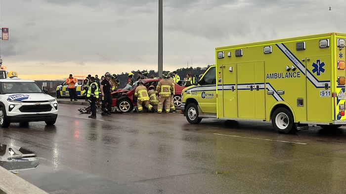 Une ambulance est stationnée près d'une voiture accidentée. Plusieurs pompiers et policiers travaillent autour.