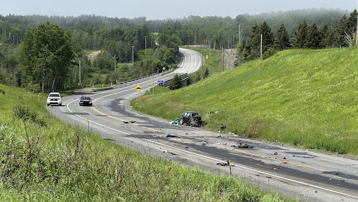 La carcasse de la voiture se trouve en bordure du chemin et des débris se trouvent sur la chaussée. Une voiture de police se trouve un peu plus loin de la scène de l'accident.