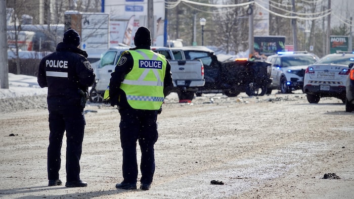 Deux policiers debout au milieu d'une route observent deux camionnettes accidentées sur le bord de la voie bloquée par des voitures de police.