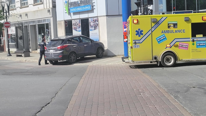 Un véhicule arrêté sur un trottoir, une ambulance tout près et une femme des services d'urgence qui arrive près du véhicule.