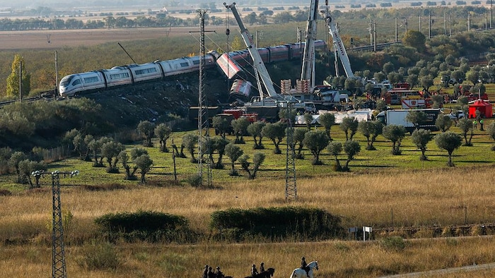 Six policiers à cheval se déplacent sur un chemin de terre dans une oliveraie qui longe le chemin de fer pendant que des grues soulèvent des wagons qui ont déraillé.