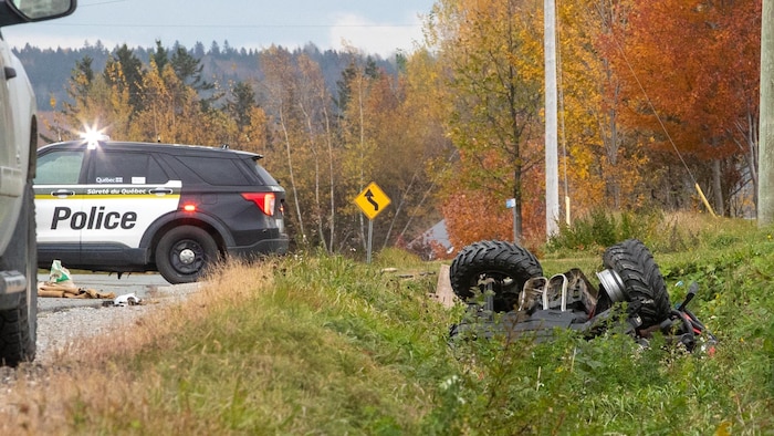 Un VTT renversé dans un fossé sur le bord d'une route avec un véhicule de police à l'arrière-plan.