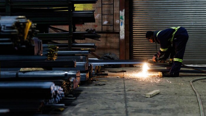 Devant des morceaux d'acier, un travailleur, penché, découpe une pièce, un processus qui fait des flammèches.