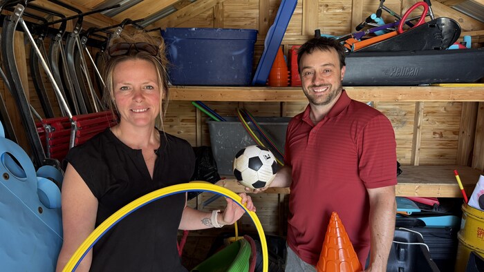 Sandy De Champlain-Tinor et Danny Pomerleau montrent notamment un cerceau, un ballon de soccer et un cône orange en prenant la pose dans un petit local de rangement.