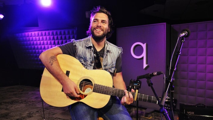 Un homme en veste de jeans sans manche par-dessus un t-shirt noir est assis dans un studio avec une guitare acoustique. Il regarde vers la gauche et sourit.
