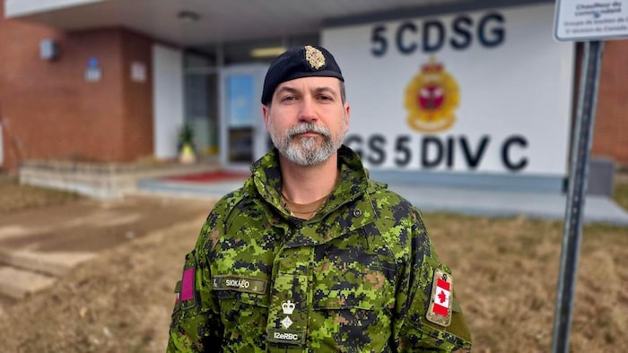 Un militaire portant une courte barbe blanche et grise, en uniforme, coiffé d'un béret, debout se tenant droit à l'extérieur devant un bâtiment de la base.