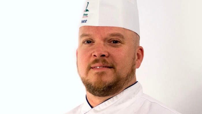 Portrait d'un chef cuisinier vêtu d'une tunique blanche et coiffé d'une très haute toque blanche, sur un fond blanc.