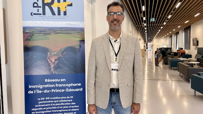 Adil Khallate pose pour la photo à côté d'une grande affiche du Réseau en immigration francophone de l'Île-du-Prince-Édouard.