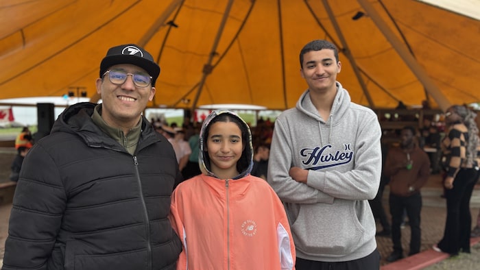 Adil Maizi with her daughter and his son, smiling. They are standing in front of a crowd of people, in a large yellow tent.