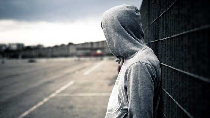 Une personne portant un chandail avec une capuche qui cache son visage est adossée à une clôture, seule.