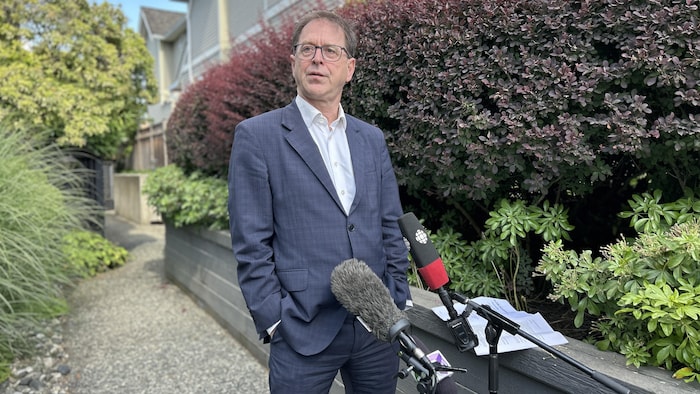 Adrian Dix est debout devant une résidence et derrière des microphones.