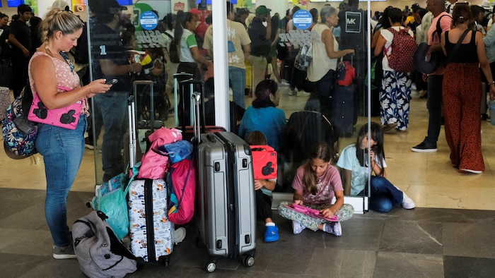 Dans un aéroport, une femme debout à côté de plusieurs valises à roulettes regarde son téléphone cellulaire, et trois enfants sont assis par terre.