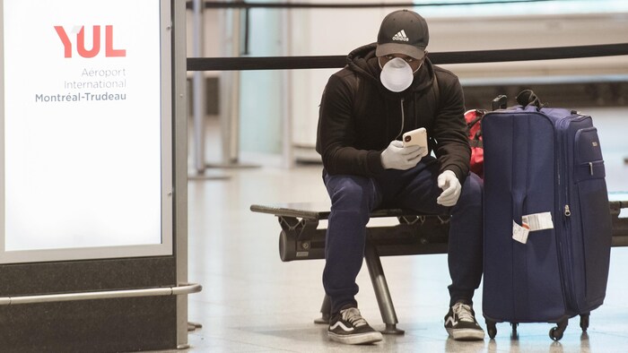 Un homme masqué et ganté attend sur un banc à côté de sa valise.