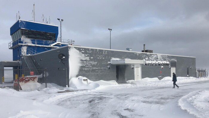 On voit la bâtisse de l'aéroport des Îles-de-la-Madeleine, en plein hiver. Une personne marche à l'extérieur et se dirige vers l'établissement.
