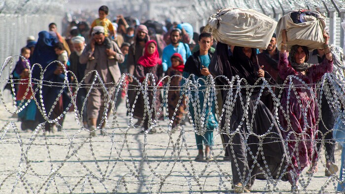 Des Afghans marchent dans un corridor protégé par du fil barbelé. Certains transportent des effets personnels. Plusieurs enfants sont visibles. 