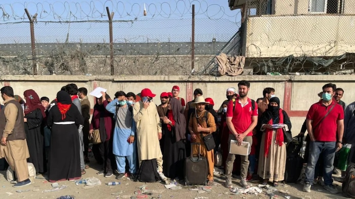 Afghan men and women with suitcases waiting to be evacuated, standing by a concrete wall with barbwire.