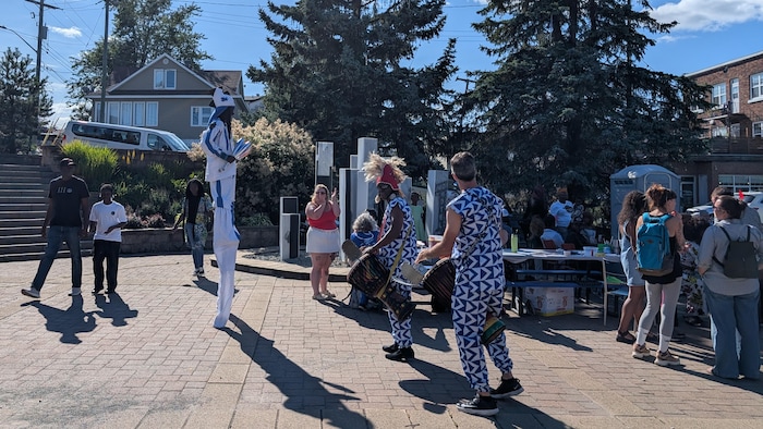 Un homme sur des échasses, à côté de deux hommes frappant sur des instruments à percussion.