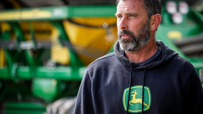 Portrait en gros plan d'un homme barbu au regard pensif, portant un chandail à capuchon noir orné du logo de la marque John Deere.