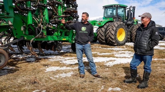 Deux hommes discutent devant une imposante machinerie agricole verte de marque John Deere dans un champ parsemé de plaques de neige.