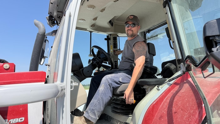 Éric Migneault, au volant de son tracteur et lunettes de soleil sur les yeux, regarde la caméra avec un léger sourire.