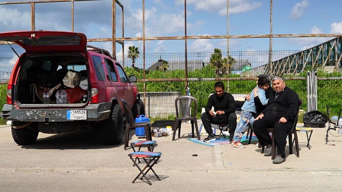 Deux hommes et une femme sont assis sur des chaises en plastique non loin d'une voiture.