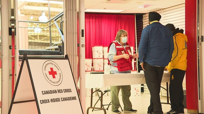 Une femme à une table portant un uniforme de la Croix-Rouge. Elle parle avec deux citoyens.
