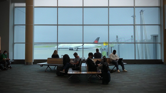 Des passagers sont assis dans une aire d'attente de l'aéroport devant une grande fenêtre. Un avion d'Air Canada est posé sur la piste.