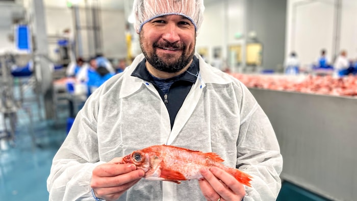 Un homme dans une usine, en combinaison protectrice blanche et bonnet. Il exhibe en souriant un poisson mort rouge.