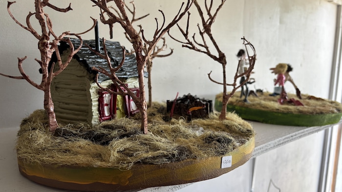 Gros plan sur une maquette sculptée montrant une cabane en bois entourée d'arbres miniatures et d'herbe séchée.